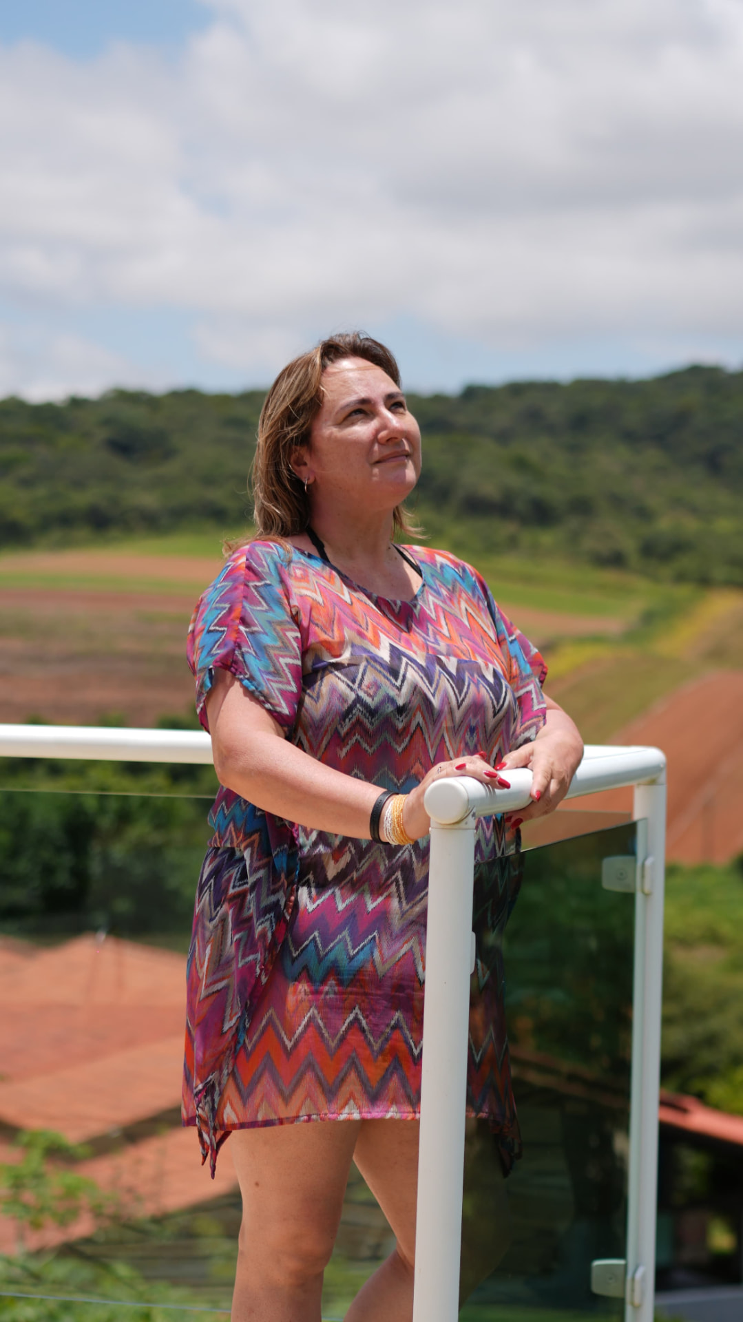 Rosi Talarico de vestido colorido, apoiada na grade, olhando o horizonte com campo verde ao fundo